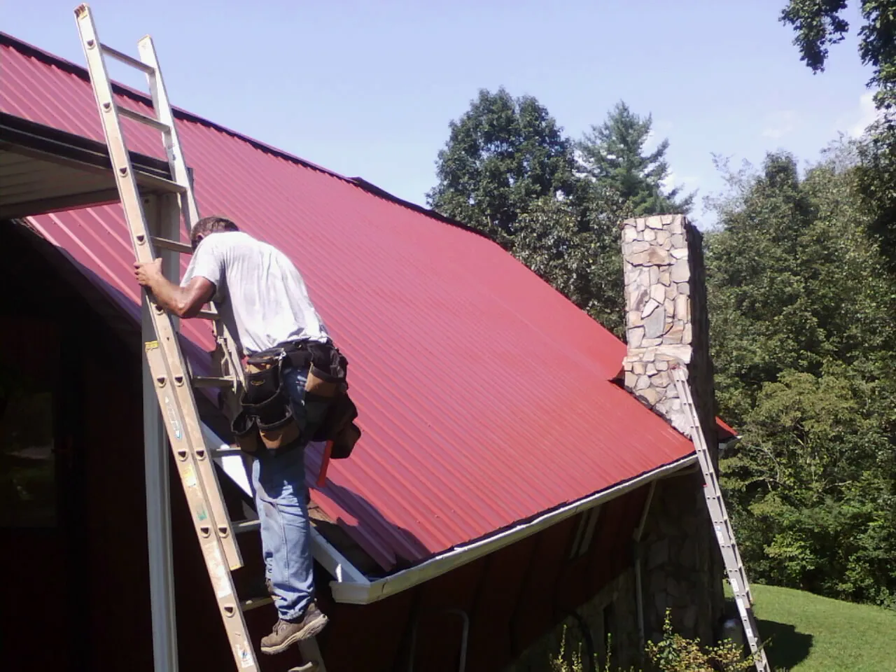Roofer on a ladder beside a red metal roof and stone chimney — Soffit & Fascia Repair by Roofing Brothers in Apollo
